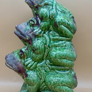 May include: A ceramic sculpture of three green frogs stacked on top of each other. The frogs have a glossy finish with some areas of dark red and gray. The base is a rectangular shape with a decorative border.