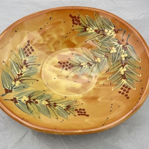 Puede incluir: Un cuenco redondo de cerámica con un esmalte cálido de color marrón anaranjado. El cuenco está decorado con hojas verdes pintadas a mano, detalles amarillos y racimos de bayas marrones. El diseño está dispuesto alrededor del borde y el centro del cuenco.