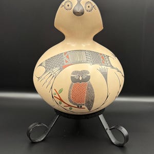Può includere: Un vaso in ceramica beige a forma di gufo con disegni geometrici neri e rossi e un motivo a gufo. Il vaso è esposto su un supporto in metallo nero con una base circolare e tre gambe curve.