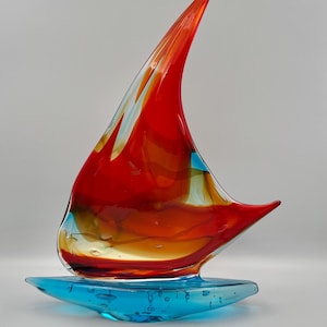 Könnte beinhalten: Eine Glasskulptur in Form eines Segelboots mit einem leuchtend roten und orangefarbenen Segel, das interne Luftblasen aufweist. Der Sockel der Skulptur ist ein massives, durchscheinendes blaues Glasstück, das das Wasser darstellt und ebenfalls kleine Luftblasen enthält.
