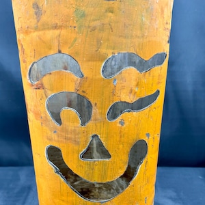 May include: A weathered, orange metal jack-o'-lantern with a carved smiling face. The cylindrical container shows signs of age and wear. The face design includes curved eye shapes, a triangular nose, and a curved mouth, typical of Halloween.