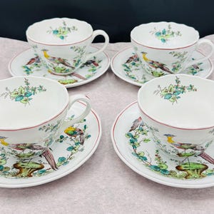 Puede incluir: Cuatro tazas y platillos blancos con un diseño de pájaros y flores. Cada taza y platillo tiene un borde rojo. El pájaro tiene plumas amarillas, marrones y moradas. También hay acentos florales verdes y azules.