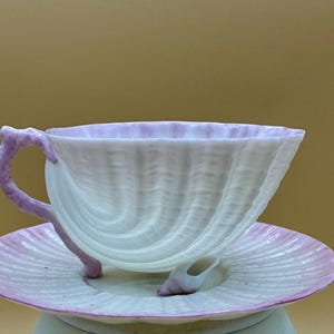 Puede incluir: Juego de taza y platillo blancos con diseño de concha. La taza y el platillo tienen una textura acanalada y están acentuados con un tono rosa pálido. El asa de la taza también es rosa. Una pequeña concha descansa sobre el platillo.