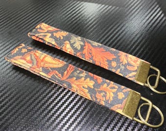 May include: Two fabric wristlet keychains featuring a dark background with a repeating pattern of orange, yellow, and brown autumn leaves. Each strap is secured with a rectangular antique brass metal clamp and a matching circular split ring.