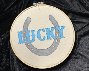 May include: A round, cream-colored embroidery hoop with a gray horseshoe design and the word "LUCKY" in blue lettering. The embroidery is set against a black background.