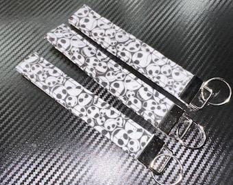 May include: Three fabric wristlet keychains featuring a repeating black and white pattern of skull faces. Each rectangular strap is finished with a silver-toned metal clamp and a circular split ring for attaching keys or other small items.