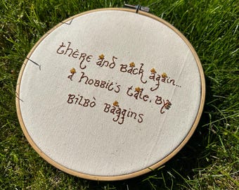 May include: A round, beige fabric embroidery hoop with brown text that reads "there and back again... a hobbit's tale, by Bilbo Baggins." The embroidery is set against a background of green grass.