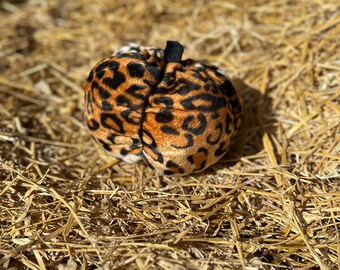 May include: A plush pumpkin with a leopard print pattern in shades of brown and black. The pumpkin has a small black stem and is set against a background of hay.
