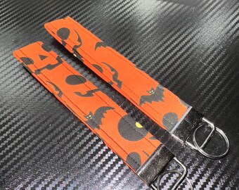 May include: Two fabric wristlet keychains featuring an orange background with a repeating pattern of black bats and black circles. Each keychain is finished with a silver-toned metal hardware clamp and a circular split ring for attaching keys.