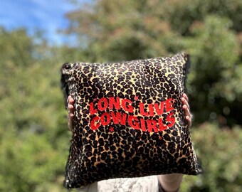 May include: Square leopard print pillow with the text "LONG LIVE COWGIRLS" in red. The pillow has a black fringe trim and is held up against a background of green foliage and a blue sky.
