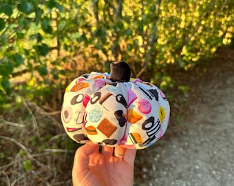 May include: A fabric pumpkin decoration featuring a white background with a colorful print of various candies and sweets, including licorice, gummies, and circular treats, topped with a small, solid black fabric stem.