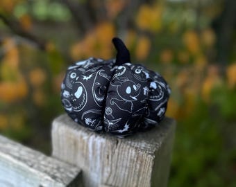 May include: A fabric pumpkin decoration featuring a black and white pattern of various character illustrations, including a ghost dog, a skeletal face, and bats. The pumpkin has a solid black fabric stem and a soft, rounded, segmented shape.