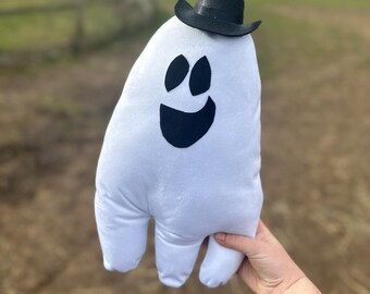 May include: A white plush ghost toy featuring a black felt face with two oval eyes and a wide smiling mouth. The ghost is wearing a small black cowboy hat perched on its head. The soft fabric body has a rounded top and three bottom points.