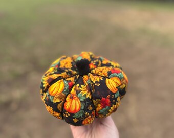 May include: A fabric pumpkin decoration featuring a black background with a colorful print of orange pumpkins, yellow sunflowers, and autumn leaves. The pumpkin has a small black fabric stem at the top and a segmented, stuffed round shape.