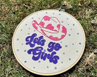 May include: An embroidered hoop art piece featuring a pink and white cowboy hat design and the phrase "Lets go Girls" in purple script. The artwork is embellished with small, sparkling accents, set against a neutral fabric background.