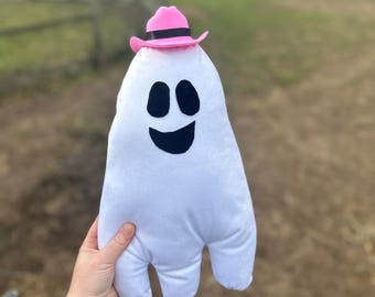 May include: A white plush ghost toy featuring a black felt face with two oval eyes and a curved mouth. The ghost is wearing a small pink cowboy hat with a black band around the crown. The toy has a rounded top and two leg-like extensions.