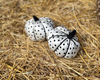 May include: Two white fabric pumpkins with black polka dots and black stems rest on a bed of hay. These decorative pumpkins are a festive fall or Halloween decoration.