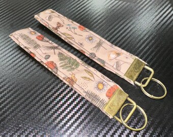 May include: Two fabric wristlet keychains featuring a light beige background with a repeating pattern of mushrooms, ferns, dragonflies, and small flowers. Each keychain is finished with a rectangular antique brass metal clamp and a round split ring.
