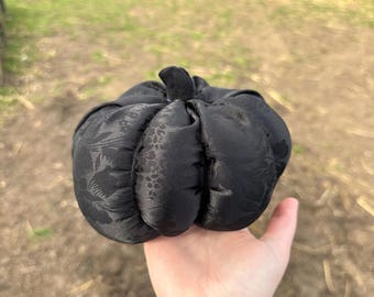 May include: A black fabric pumpkin with a subtle tonal floral pattern and a small stem at the top. The pumpkin has a segmented, rounded shape with visible stitching between the sections, creating a soft, stuffed appearance throughout the object.