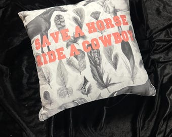 May include: Square decorative pillow with a white background featuring grayscale horse and feather illustrations. The pillow has the text "SAVE A HORSE RIDE A COWBOY" in coral-colored letters.