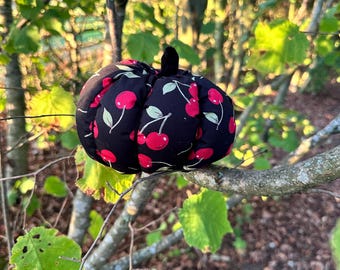 May include: A stuffed fabric pumpkin decoration featuring a black background with a repeating pattern of red cherries and green leaves. The pumpkin has a rounded, segmented shape with a small, dark, upright stem at the top center of the object.