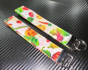 May include: Two fabric wristlet keychains featuring a white background with a colorful pattern of various vegetables including red tomatoes, orange carrots, green leafy greens, and yellow squash, each finished with a silver metal clasp and ring.