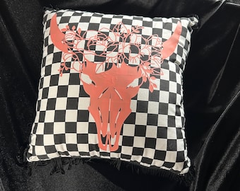 May include: Square decorative pillow with a black and white checkered pattern. The pillow features a coral-colored bull skull design with floral accents. The edges of the pillow have a fringed trim, adding a rustic touch to the design.