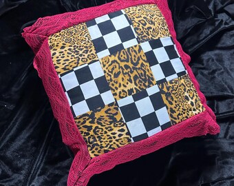 May include: Square decorative pillow with a patchwork design. The center features alternating squares of leopard print and black and white checkered patterns. The pillow is trimmed with a red lace border.