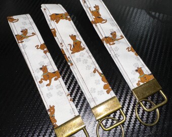 May include: Three white wrist keychains with a repeating Scooby-Doo pattern in brown and tan. Each keychain has a gold-colored metal clasp and ring. The keychains are displayed on a dark, textured surface.