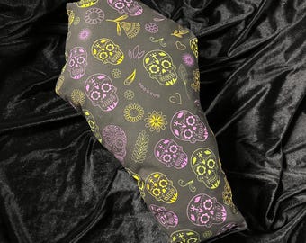 May include: A black coffin-shaped pillow with a repeating pattern of sugar skulls in yellow, pink, and green, along with floral and leaf accents. The pillow is resting on a black velvet surface.