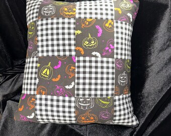 May include: Square decorative pillow with a patchwork design. The pillow features alternating squares of black and white gingham and a dark background with Halloween-themed illustrations of jack-o'-lanterns and bats in orange, purple, and yellow.