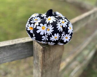 May include: A fabric pumpkin decoration featuring a black background with a repeating white daisy pattern and yellow centers. The pumpkin has a dark, solid-colored stem at the top and a rounded, segmented shape with soft, stuffed fabric construction.