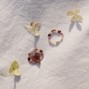 May include: Two beaded bracelets on a white fabric background. One bracelet is a simple circle of pink and white beads. The other bracelet is a heart shape with pink and white beads.