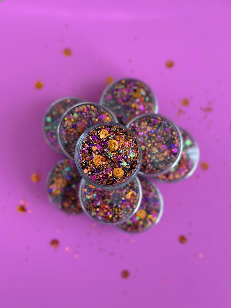 May include: A collection of small, clear containers filled with colorful glitter. The glitter is a mix of orange, purple, and green, with some small pumpkin-shaped confetti. The containers are arranged on a bright purple background.
