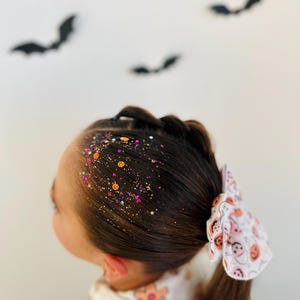 May include: A young person's dark brown hair styled with a side braid and ponytail, adorned with colorful glitter and a white bow featuring a Halloween-themed pattern. Black bat silhouettes are in the background.