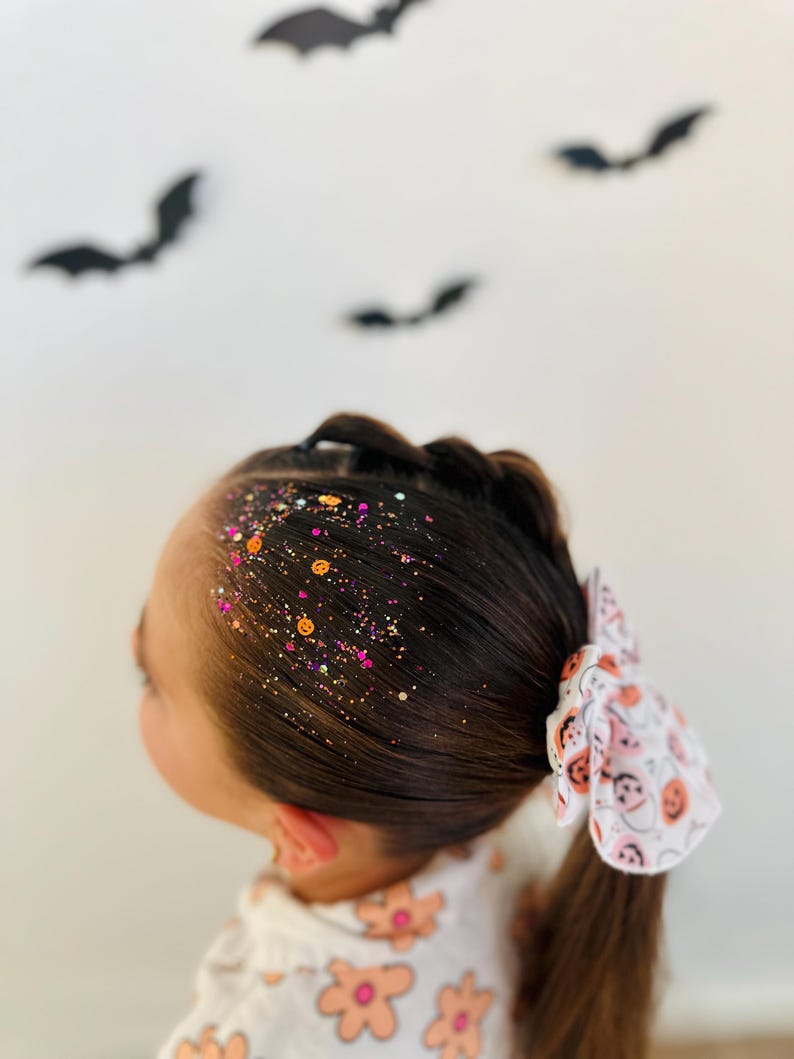 May include: A young person's dark brown hair styled with a side braid and ponytail, adorned with colorful glitter and a white bow featuring a Halloween-themed pattern. Black bat silhouettes are in the background.
