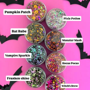 May include: A collection of eight small, clear containers filled with colorful glitter mixes, each labeled with a Halloween-themed name like "Pumpkin Patch" and "Witch's Brew." Black bat silhouettes decorate the bright pink background.