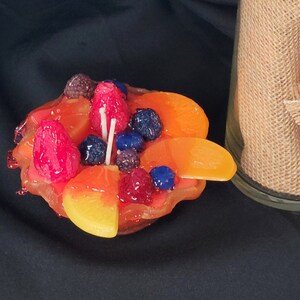 May include: A fruit-shaped candle with a yellow, orange, and red design. The candle is decorated with blueberries, raspberries, and blackberries. The candle is sitting on a black surface.