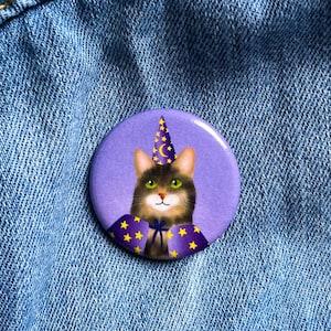 Puede incluir: Un botón redondo morado con un gato con sombrero y capa de mago con estrellas doradas. El gato tiene ojos verdes y hocico blanco. El botón está prendido a una chaqueta vaquera azul.