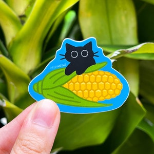 May include: A sticker depicting a black cat on a yellow corn cob with green leaves. The sticker has a blue outline and the text "crispymeows" is printed on the bottom left.