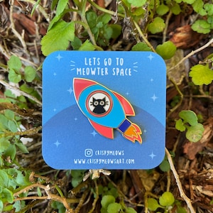 Puede incluir: Un pin de esmalte azul con un cohete con un gato negro en el centro. El cohete tiene detalles en rojo y azul con llamas naranjas. El texto en el pin dice "Lets go to Meowter Space" e incluye Instagram y el sitio web de Crispy Meows.
