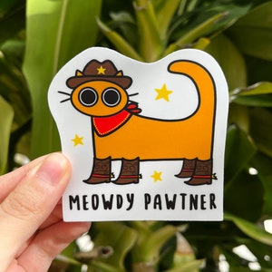 May include: A sticker depicting an orange cartoon cat wearing a cowboy hat, red bandana, and cowboy boots. The text "MEOWDY PAWTNER" is displayed below the cat, with yellow star accents. The cat has large, round eyes.