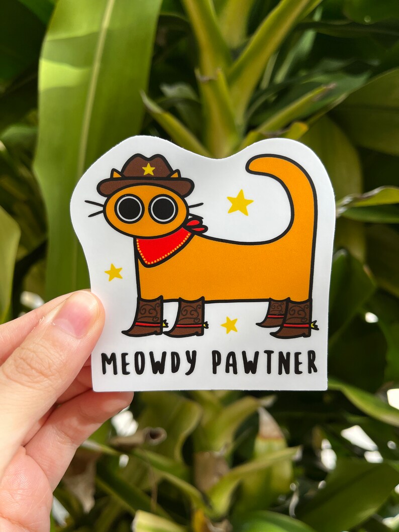 May include: A sticker depicting an orange cartoon cat wearing a cowboy hat, red bandana, and cowboy boots. The text "MEOWDY PAWTNER" is displayed below the cat, with yellow star accents. The cat has large, round eyes.