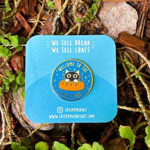 Puede incluir: Una tarjeta azul con el texto "WE SELL BREAD WE SELL LOAFS" y un pin con un gato negro asomándose por encima de una barra de pan. El pin dice "WELCOME TO THE BREAD BANK". La tarjeta también incluye la dirección de Instagram y el sitio web del artista.