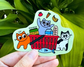 Puede incluir: Una pegatina con gatos de dibujos animados con una cesta de la compra llena de comestibles. La pegatina incluye una cesta roja, un pez azul, una barra de pan y un plátano. El texto "CRISPYMEOWS" también es visible.