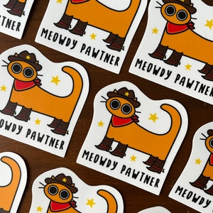 May include: Stickers with a cartoon cat design. The cat is orange with a cowboy hat, red bandana, and boots. The text "MEOWDY PAWTNER" is printed below the cat. The stickers are on a wooden surface.