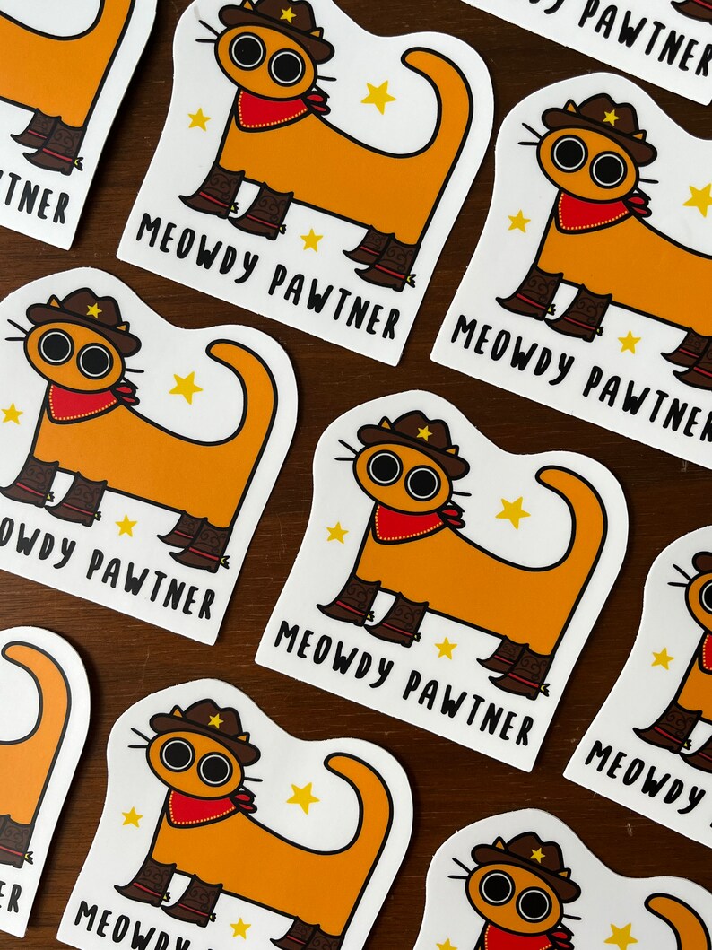 May include: Stickers with a cartoon cat design. The cat is orange with a cowboy hat, red bandana, and boots. The text "MEOWDY PAWTNER" is printed below the cat. The stickers are on a wooden surface.