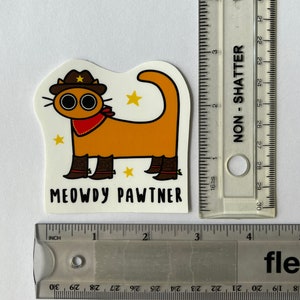 May include: A sticker depicting an orange cartoon cat wearing a cowboy hat, boots, and a red bandana. The sticker has the text "MEOWDY PAWTNER". The sticker is approximately 7.6 cm in length.