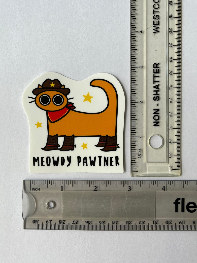 May include: A sticker depicting an orange cartoon cat wearing a cowboy hat, boots, and a red bandana. The sticker has the text "MEOWDY PAWTNER". The sticker is approximately 7.6 cm in length.
