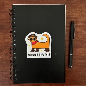 May include: A black spiral-bound notebook with a sticker of an orange cat wearing a cowboy hat and the text "MEOWDY PAWTNER." A black pen rests on a wooden surface next to the notebook.